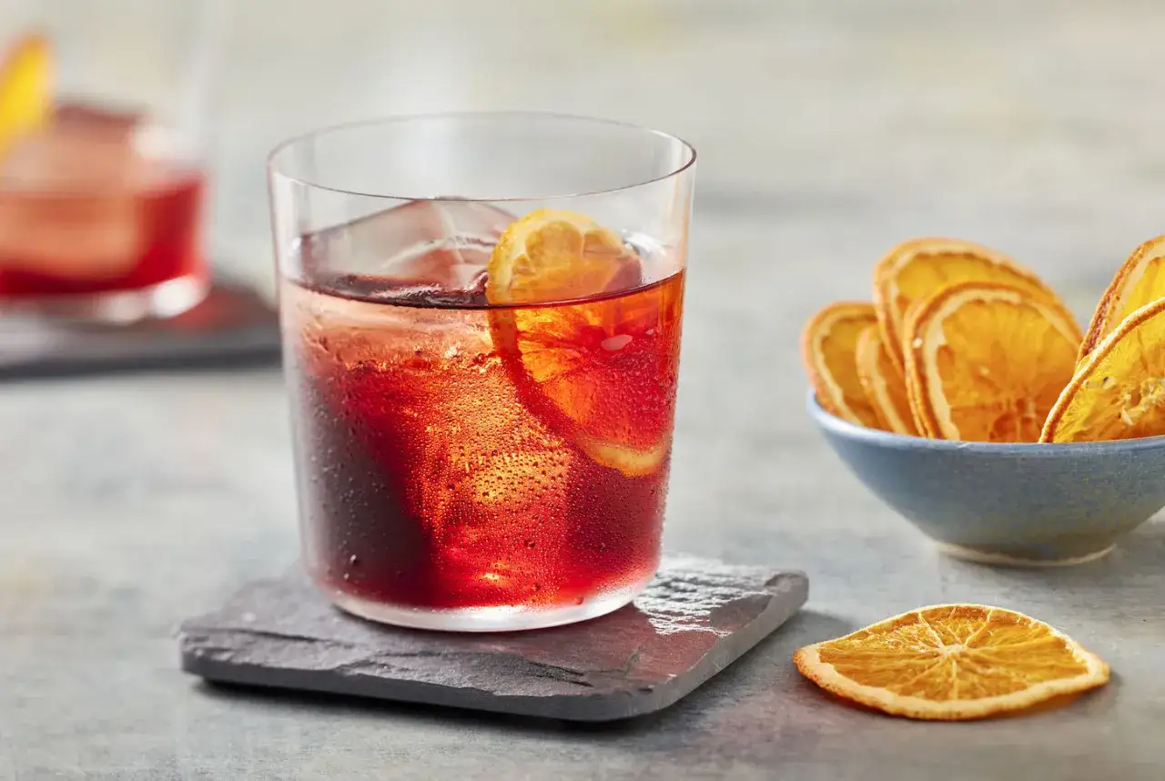 Ein erfrischender Negroni, das negroni rezept original mit Eis und Orangenscheibe. Daneben eine Schale mit getrockneten Orangen.