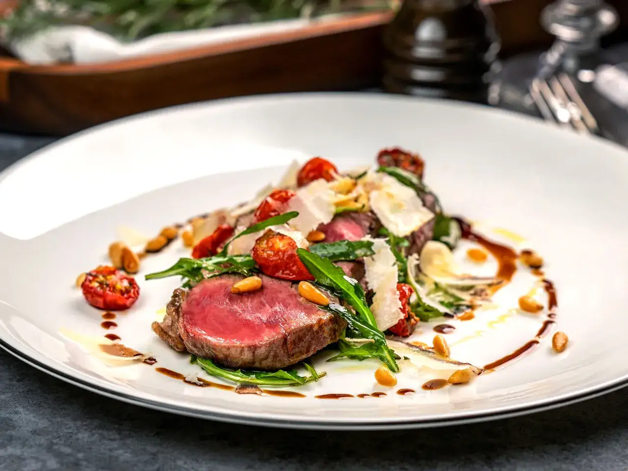 Saftiges Tagliata di manzo mit Rucola, Kirschtomaten, Pinienkernen und Parmesan auf einem weißen Teller.