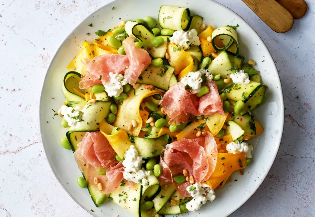 Ein bunter, sommerlicher Zucchini-Salat mit Edamame, Pinienkernen, Prosciutto und Frischk&auml;se auf einem Teller.