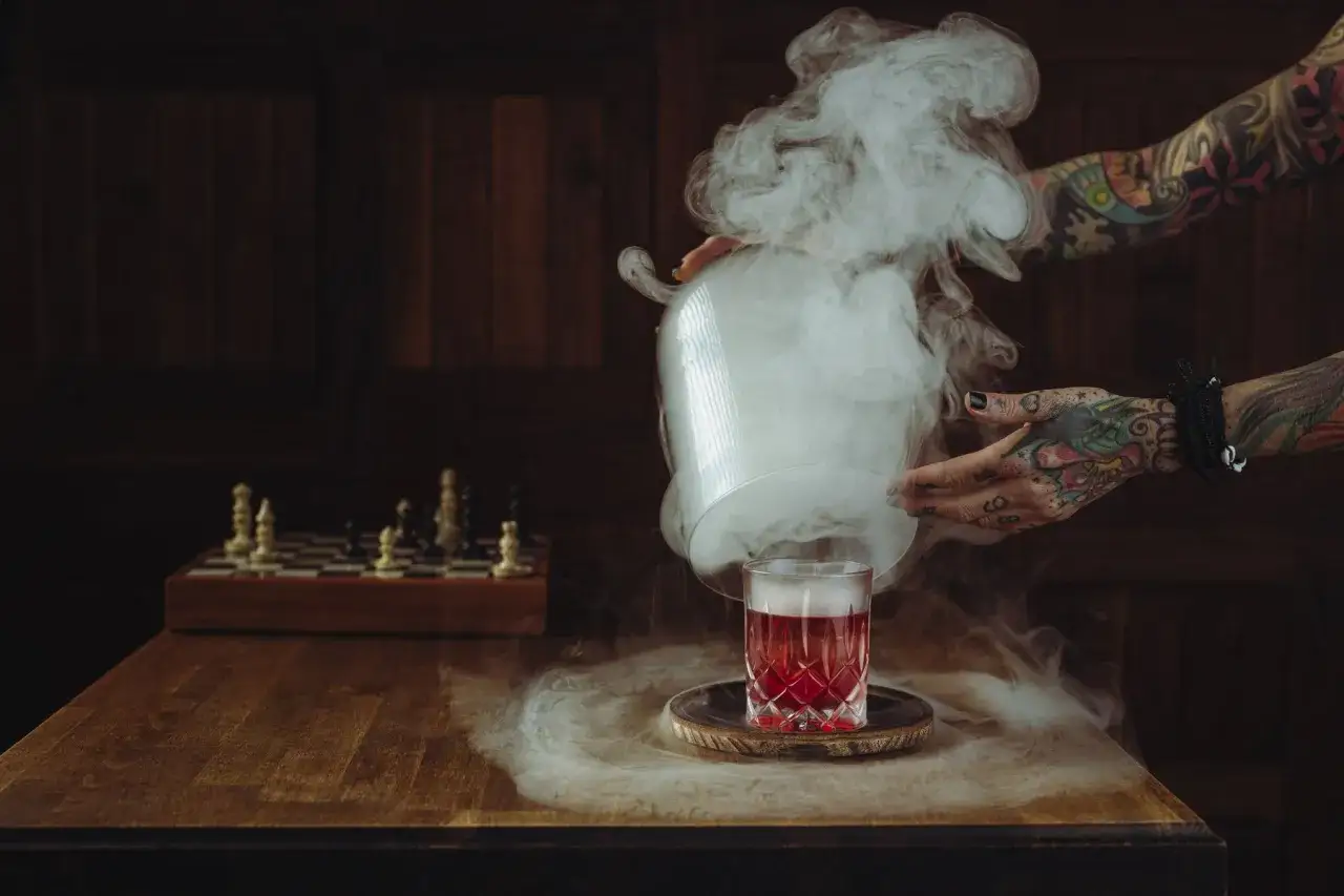 Ein Glas mit rotem Wermut, umgeben von Rauch. Ein Schachbrett steht im Hintergrund.