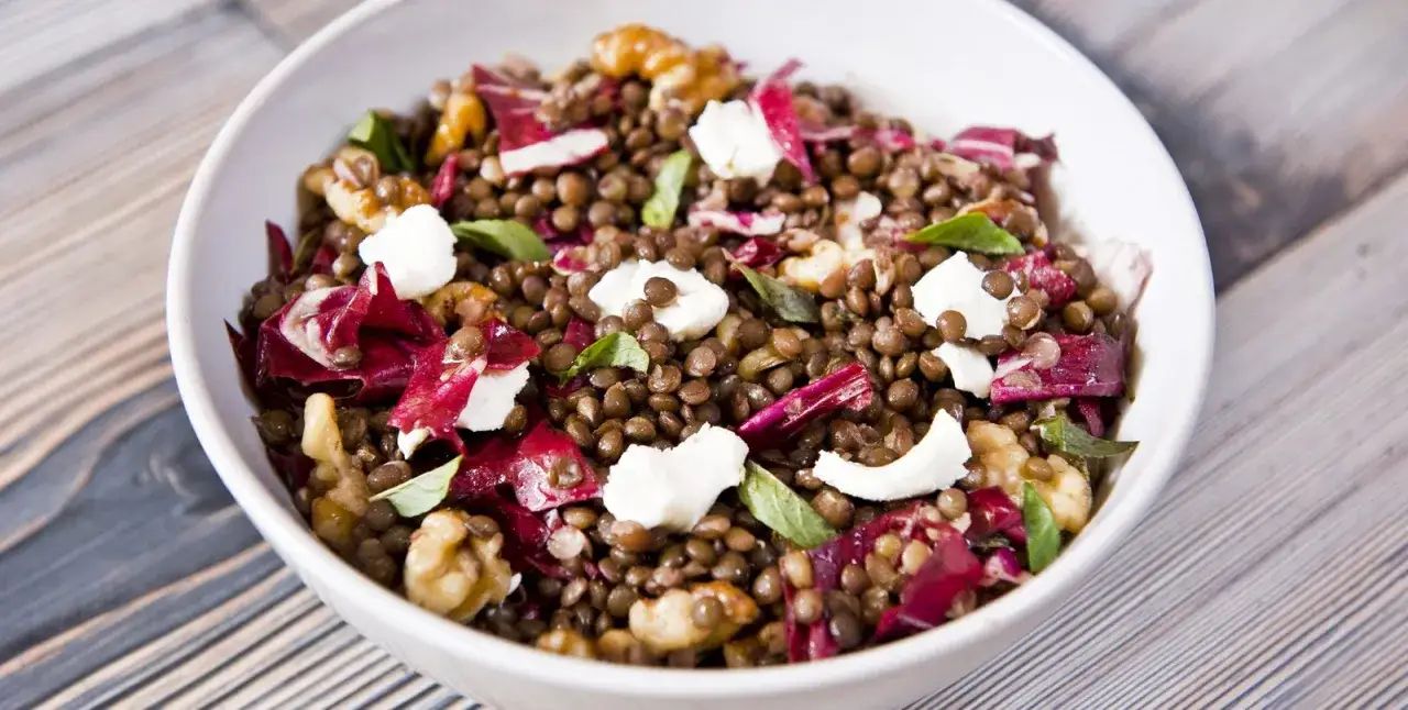 Linsensalat mit Feta und Paprika, garniert mit Waln&uuml;ssen und frischen Kr&auml;utern, in einer wei&szlig;en Sch&uuml;ssel auf Holzhintergrund.