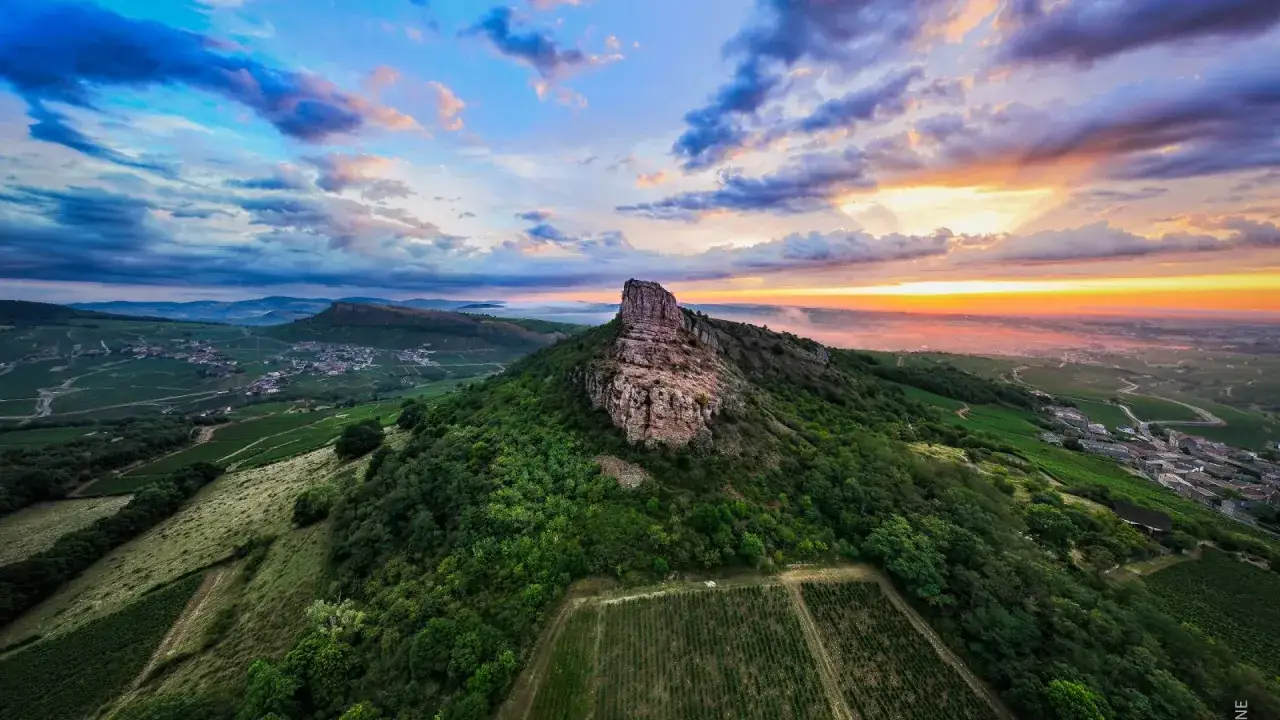 Sonnenuntergang &uuml;ber den Weinbergen von Pouilly-Fuiss&eacute;. Ein steiler Felsen ragt aus einem gr&uuml;nen H&uuml;gel, umgeben von Reben und einem Dorf.