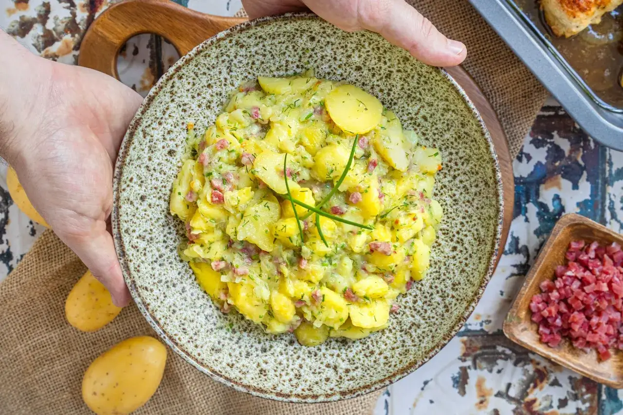 Ein Teller mit Kartoffelsalat mit Speck, garniert mit Schnittlauch. Daneben liegen rohe Kartoffeln und eine Schale mit gew&uuml;rfeltem Speck.