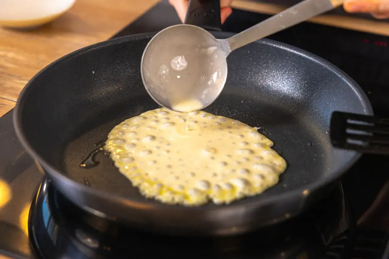 Quarkpfannkuchen werden in der Pfanne gebacken. Ein Schöpflöffel gibt Teig hinein.