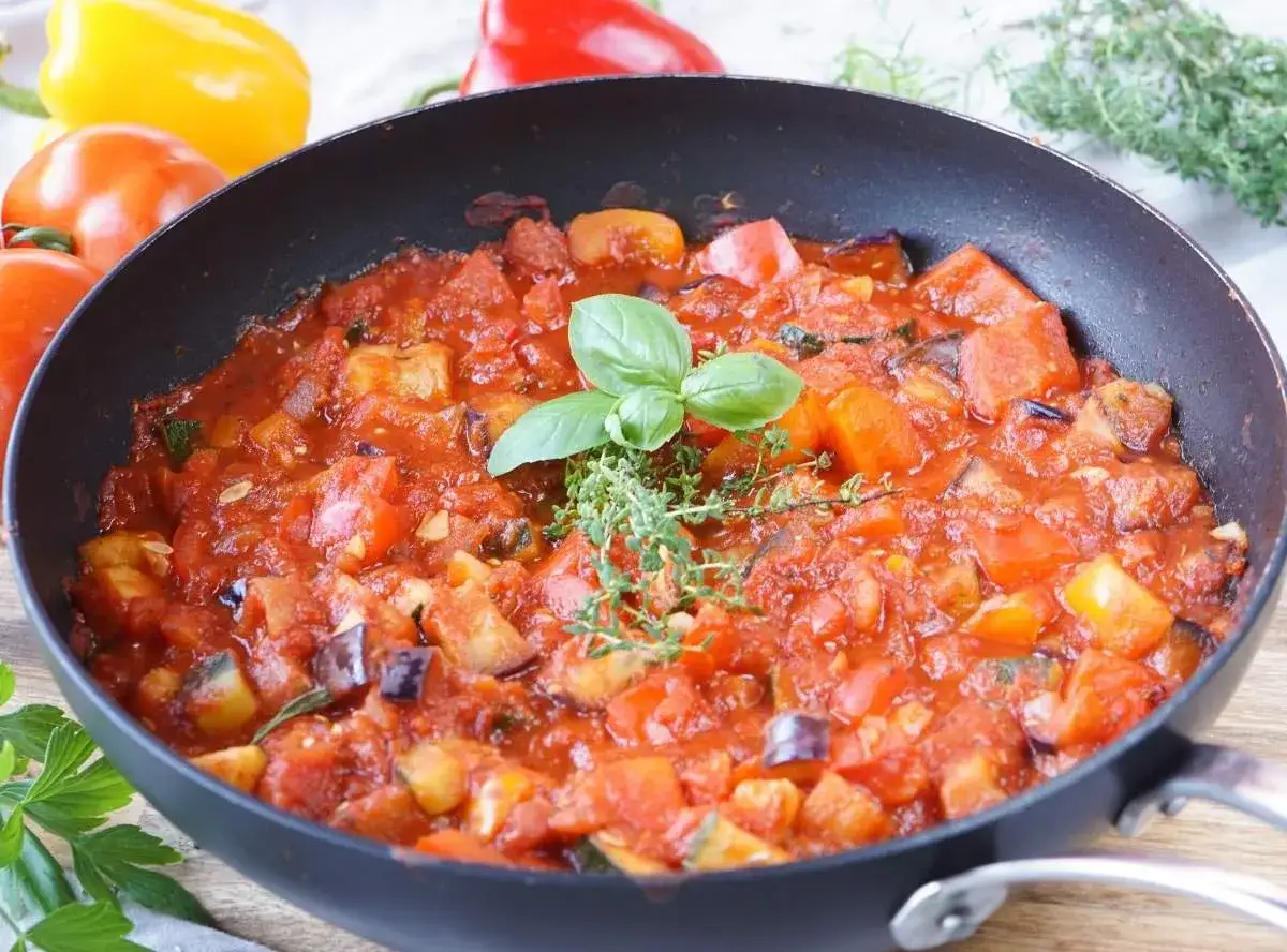 Ein k&ouml;stliches Ratatouille-Rezept in der Pfanne, garniert mit frischem Basilikum und Thymian. Bunte Paprika im Hintergrund.