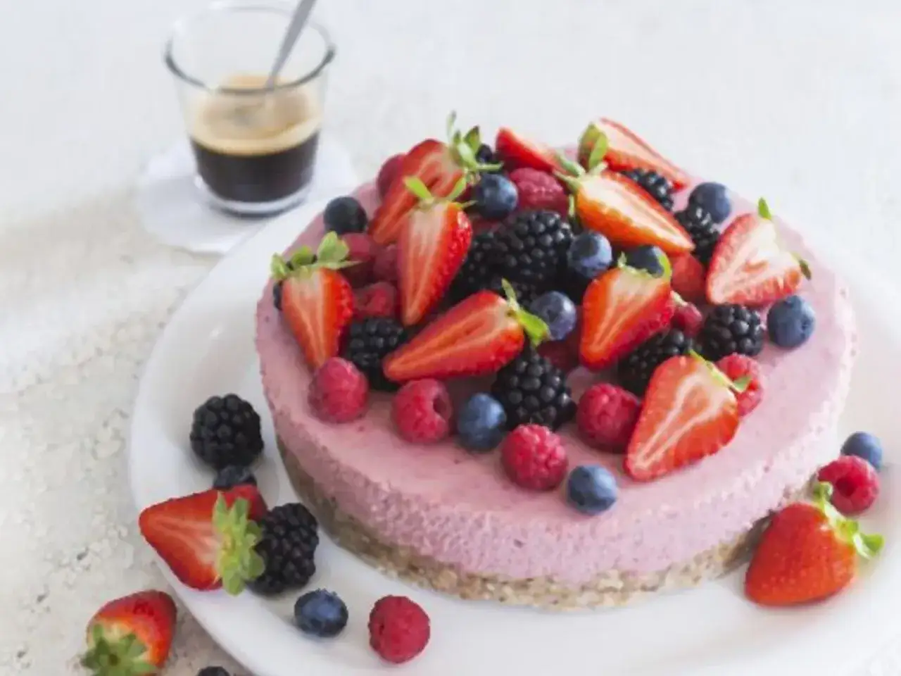 Eine schnelle Torte mit frischen Beeren wie Erdbeeren, Blaubeeren und Himbeeren, garniert auf einer cremigen rosa Schicht. Daneben ein Glas Espresso.