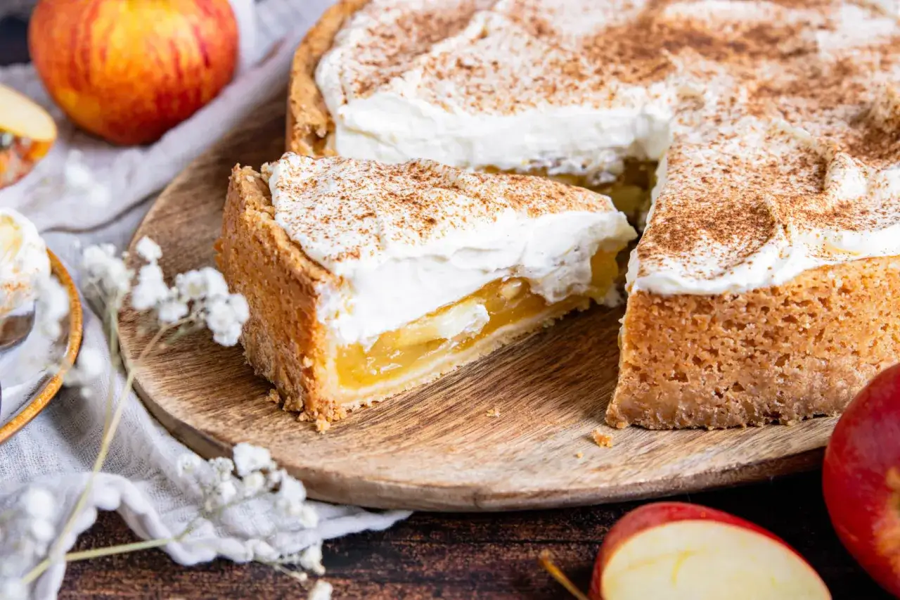 Ein Stück Apfel Schmand Torte mit cremigem Topping und Zimt bestreut. Daneben liegen frische Äpfel.
