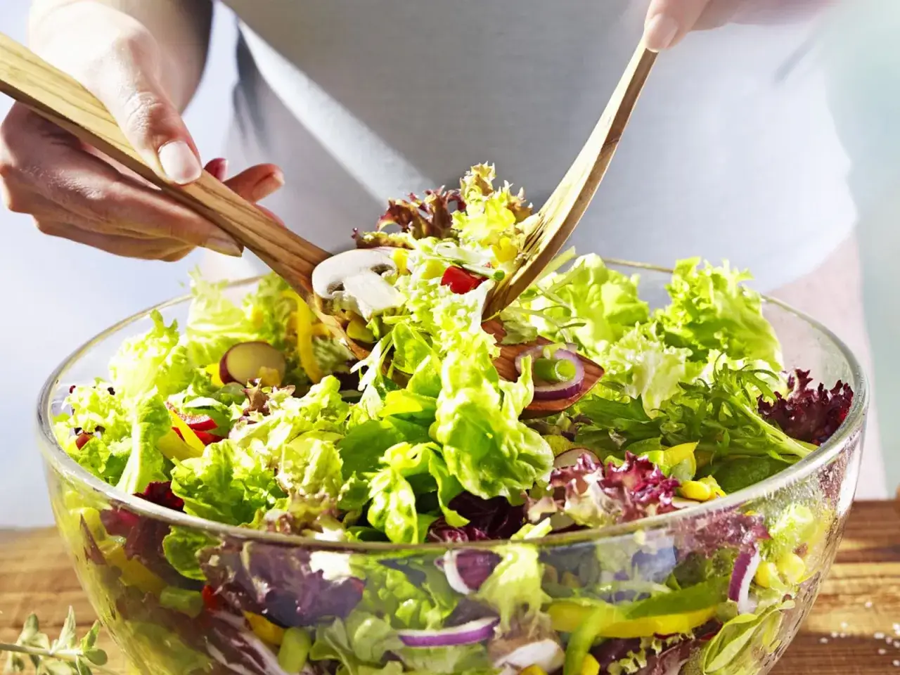 H&auml;nde mischen mit Holzschaufeln einen bunten gemischten Salat in einer Glassch&uuml;ssel. Frische Zutaten wie Radieschen, Zwiebeln und Pilze sind sichtbar.