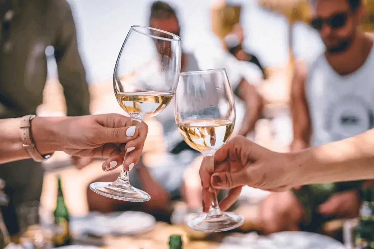 Zwei H&auml;nde sto&szlig;en mit Gl&auml;sern an, gef&uuml;llt mit hellem, leicht rauchigem Wein. Im Hintergrund sitzen Freunde in entspannter Runde.