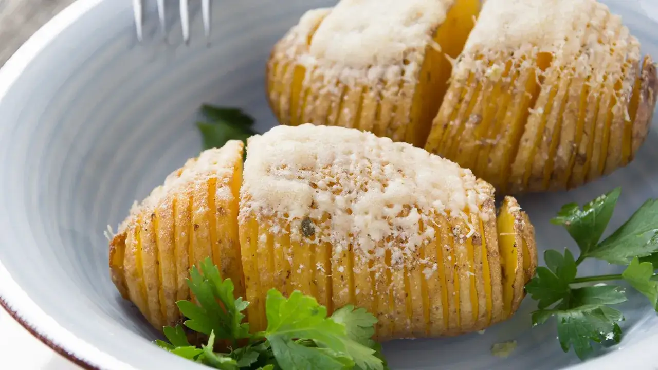 Zwei goldbraune Hasselback Kartoffeln mit geschmolzenem Käse und Petersilie in einer blauen Schüssel.