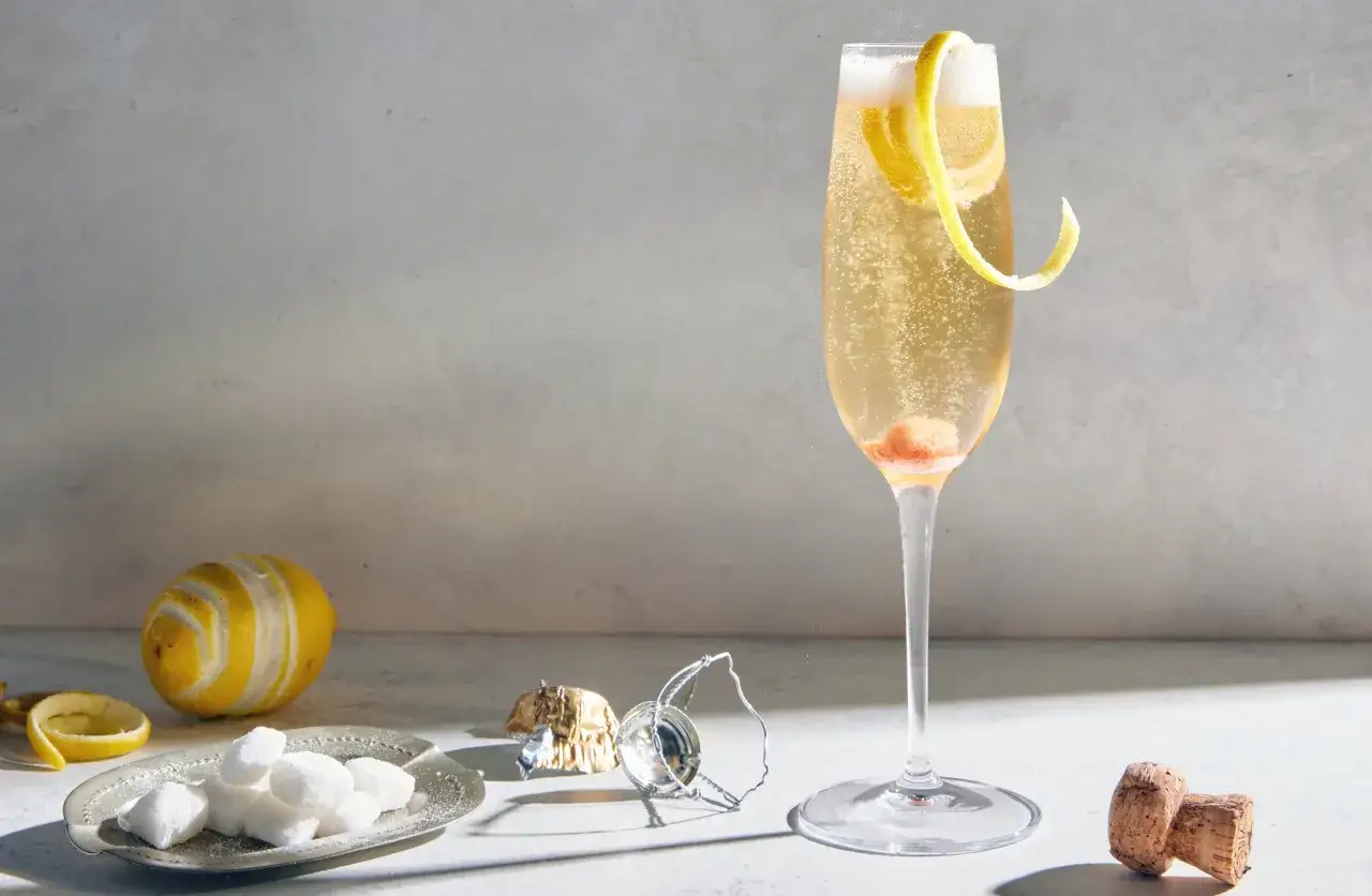 Ein prickelnder Champagner Cocktail mit Zitronenzeste und einem Hauch von Frucht. Daneben liegen Zuckerw&uuml;rfel, eine Zitrone und ein Korken.