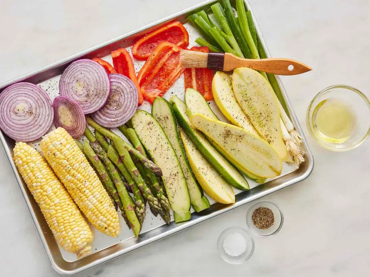 Vorbereitung für einen schnellen Salat zum Grillen: Maiskolben, Spargel, Zucchini, rote Zwiebeln und Paprika auf einem Blech, bereit zum Würzen.
