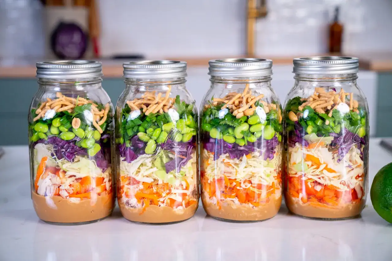 Vier Gl&auml;ser mit bunten Schichten: Erdnussdressing, Karotten, Wei&szlig;kohl, Rotkohl, Edamame, Fr&uuml;hlingszwiebeln und knusprige Nudeln. Perfekt als salate zum vorbereiten am vortag.