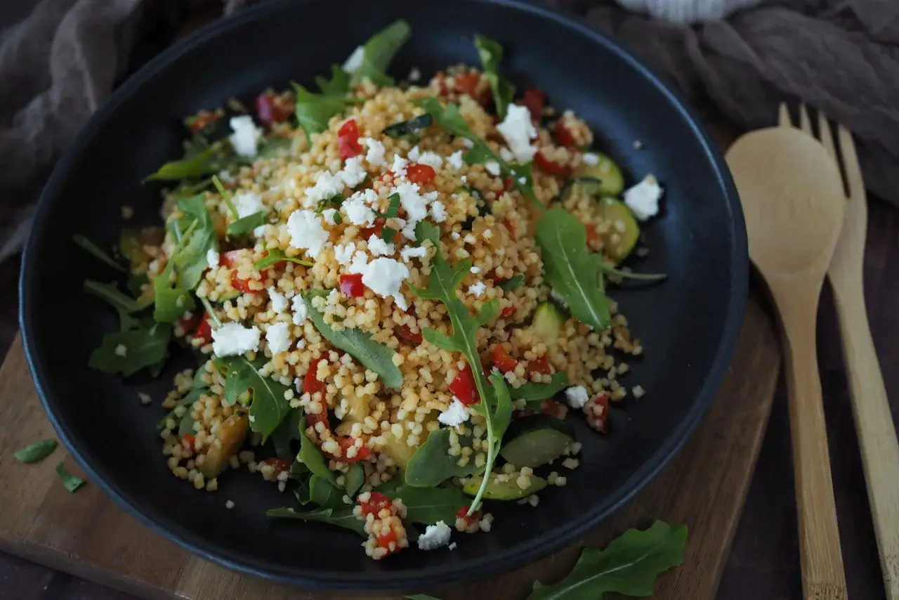 Ein bunter Couscous-Salat mit Feta, Rucola, Tomaten und Zucchini, serviert in einer schwarzen Sch&uuml;ssel.