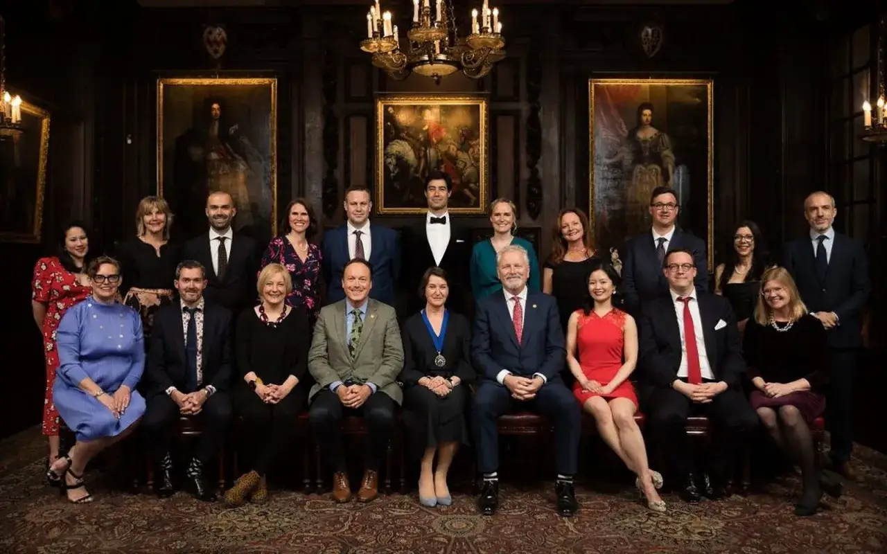 Gruppe von Personen in festlicher Kleidung, darunter ein Master of Wine, posiert f&uuml;r ein Gruppenfoto in einem eleganten Raum mit Gem&auml;lden und Kronleuchtern.