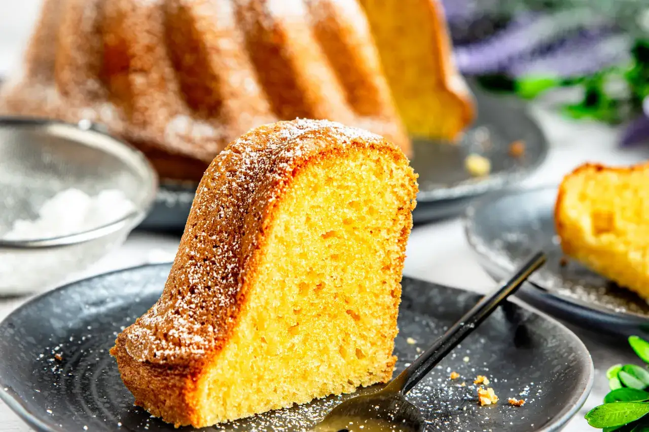Ein saftiges St&uuml;ck Paradiescreme Kuchen, best&auml;ubt mit Puderzucker, liegt auf einem schwarzen Teller.