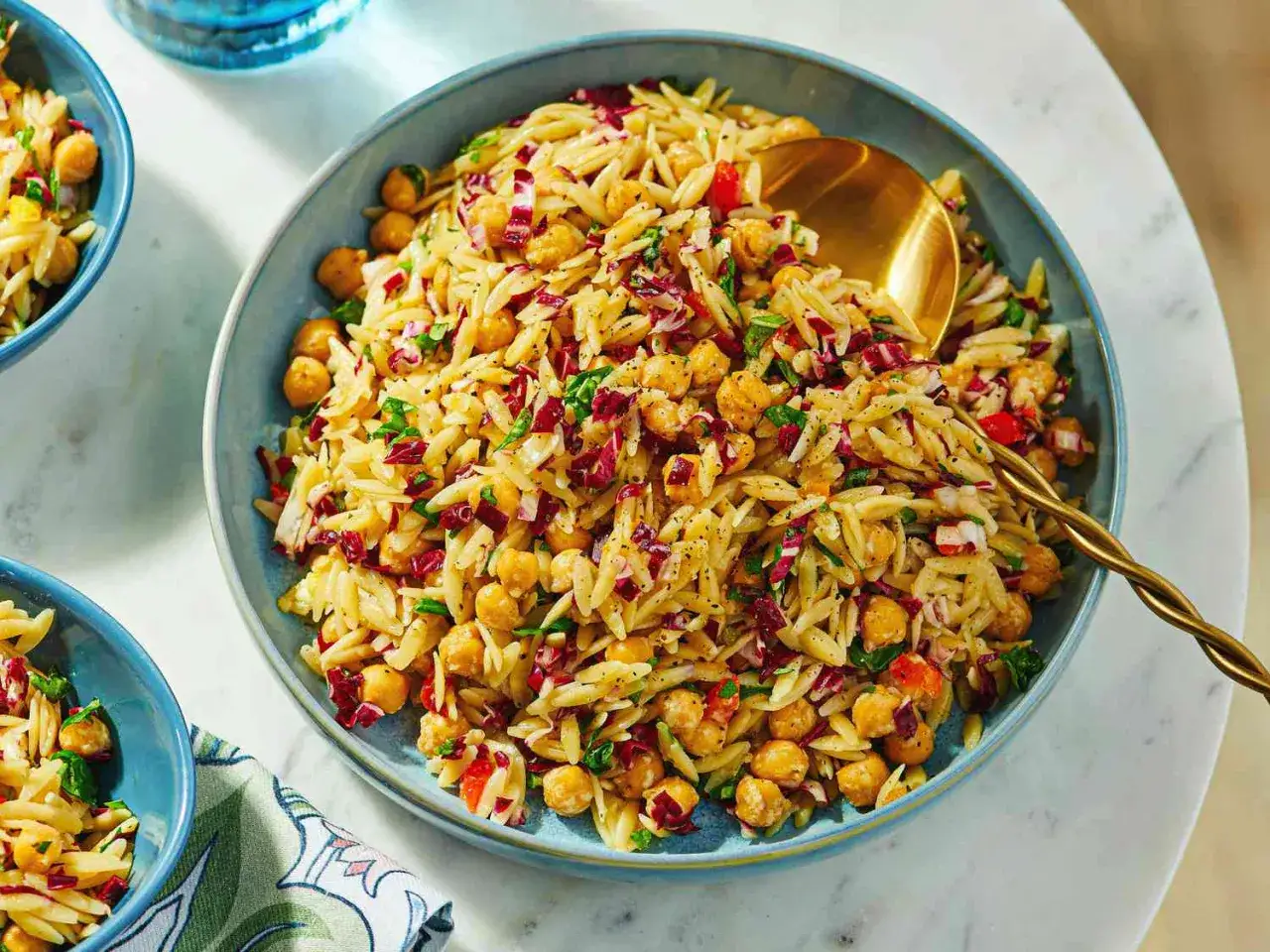 Ein bunter Nudelsalat mal anders: Orzo-Nudeln mit Kichererbsen, Radicchio und Kr&auml;utern in einer blauen Sch&uuml;ssel, serviert mit einem goldenen L&ouml;ffel.