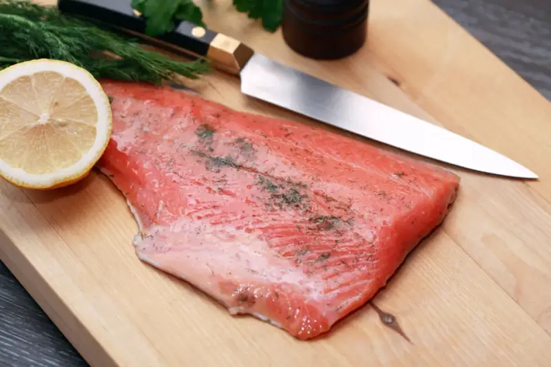Frisches Lachsfilet mit Dill und Zitrone auf einem Holzbrett, bereit zum Kochen mit Gin. Ein scharfes Messer liegt daneben.